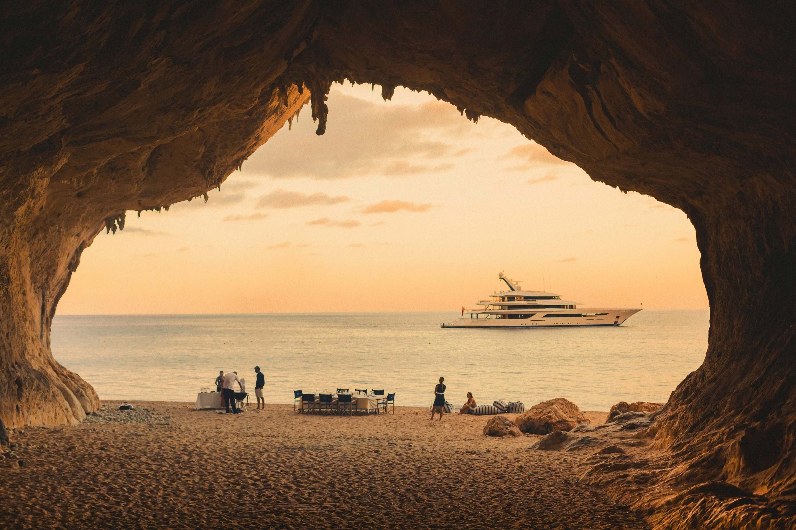 Inside a cave, a yacht is anchored as beachgoers enjoy the sun on the shore.
