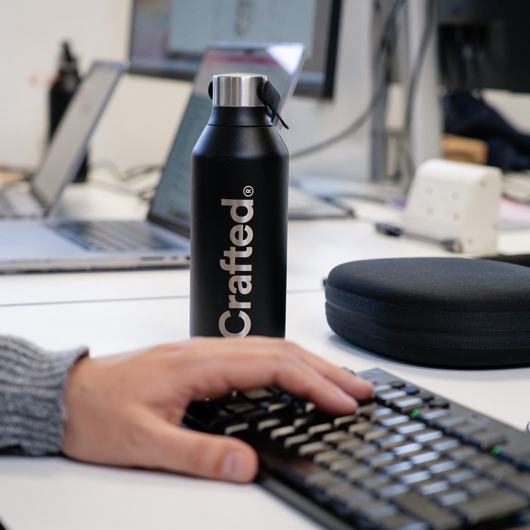 A person typing on a keyboard, hands positioned over the keys. The focal point of the image is a black water bottle with 'Crafted' engraved on the side.
