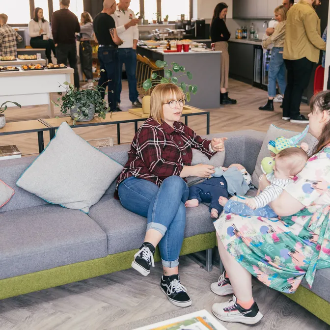 Two women with babies sit together on a couch in a modern office setting, engaged in conversation.